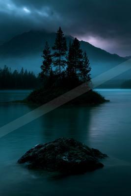 A small rocky island covered in tall evergreen trees sits in the middle of a calm mountain lake, with a large dark rock in the foreground and misty mountains un
