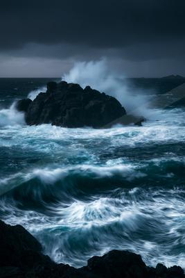 Powerful ocean waves with white foam crashing against a large dark rock formation under a stormy sky, creating a dramatic seascape.