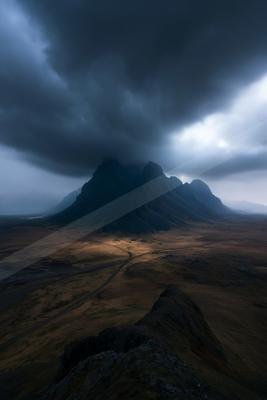 Moody atmospheric landscape with dark storm clouds enveloping a jagged mountain peak, with light breaking through and casting rays on the barren valley below.