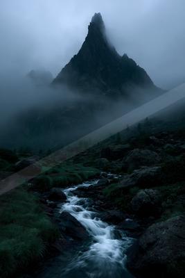 A dramatic, jagged mountain peak rises through thick fog above a rocky stream with white water flowing through a green, boulder-strewn valley.