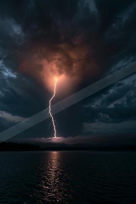 A powerful lightning bolt strikes from a stormy sky into a dark body of water, illuminating the clouds with an orange glow and reflecting on the rippled surface