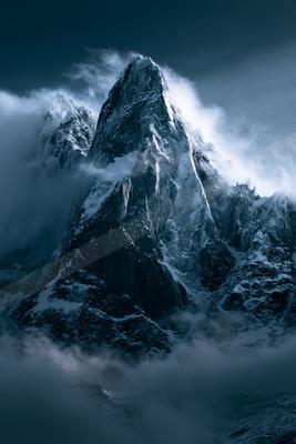 A sharp, snow-covered mountain peak rises dramatically through thick white clouds against a dark blue sky.
