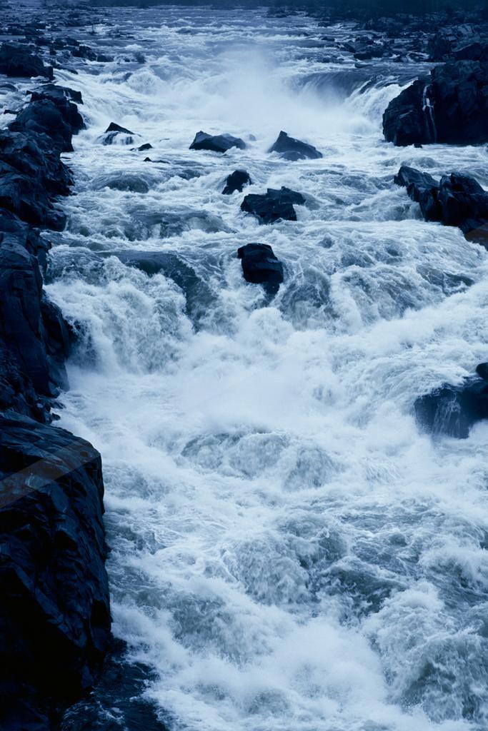 Turbulent whitewater rapids cascading forcefully over large dark rocks in a powerful river, creating mist and foam.