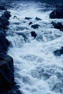 Turbulent whitewater rapids cascading forcefully over large dark rocks in a powerful river, creating mist and foam.