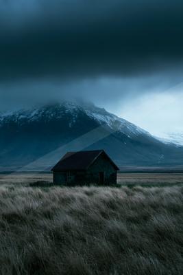 A weathered stone house stands alone in a field of dry grass under a dark, cloudy sky with a large snow-dusted mountain in the background.