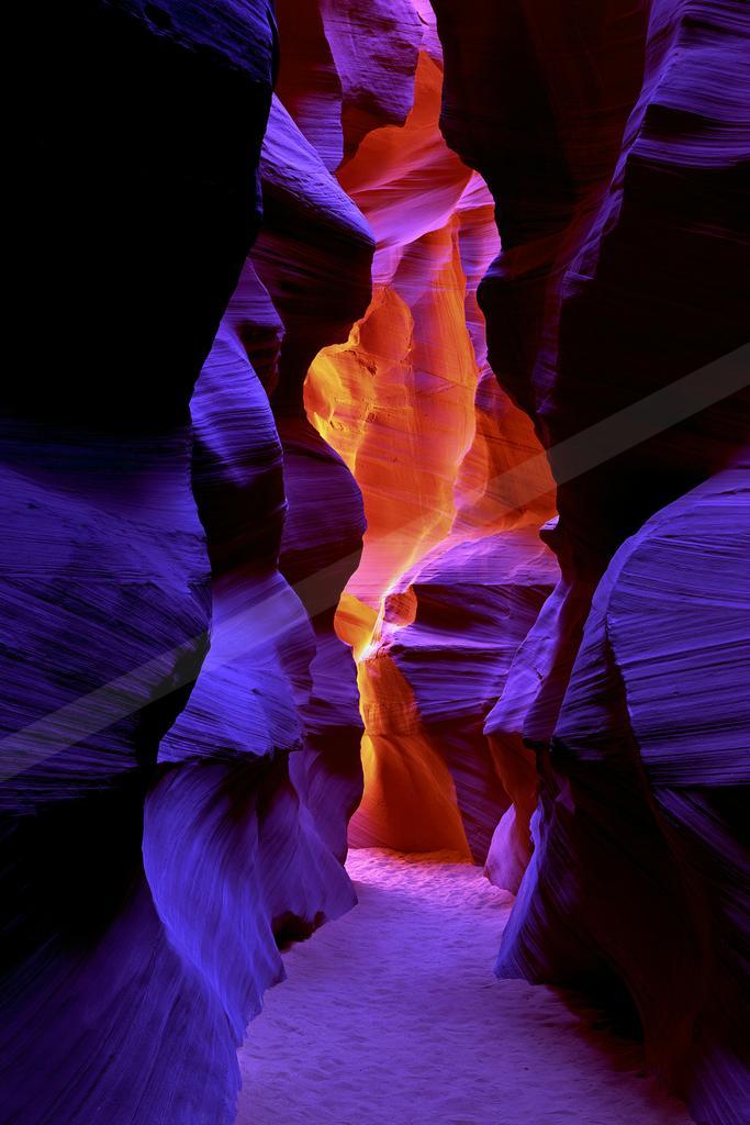 A narrow slot canyon with smooth, wavy sandstone walls illuminated by dramatic beams of orange and purple light filtering from above.