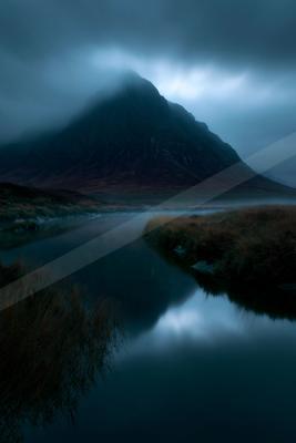 A calm lake reflects a large, dark mountain partially shrouded in thick fog and low clouds under a moody blue sky.