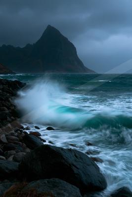 Powerful turquoise and white waves crash violently against a dark rocky shoreline, creating a large spray of water, with a steep mountain rising in the backgrou