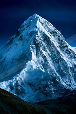 A dramatic, snow-covered mountain peak rises sharply against a deep blue twilight sky, with rocky ridges and crevasses visible on its steep slopes.