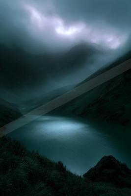A serene mountain lake surrounded by steep, fog-covered hills under a dramatic, cloudy sky with a soft glow breaking through the mist.