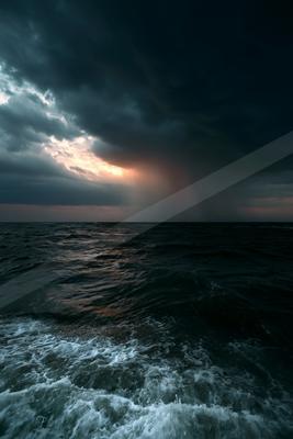 Dark stormy ocean with white-capped waves under heavy rain clouds at dusk, with a break in the clouds revealing warm light on the horizon.