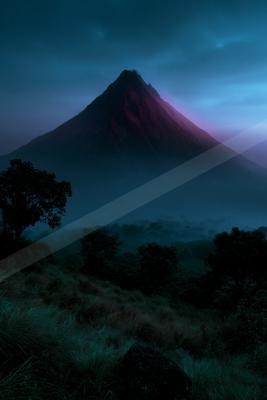 A large conical volcano rises dramatically into a dark blue twilight sky, its slopes illuminated with a pinkish glow, surrounded by thick fog and silhouetted tr