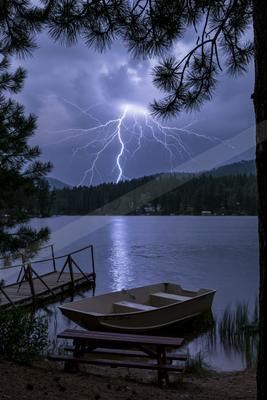Dramatic lightning bolt strikes over a mountain lake during a thunderstorm, with a small boat, wooden dock, picnic table, and pine tree branches in the foregrou