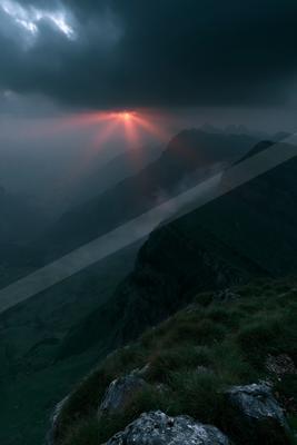 A dramatic red sun breaks through heavy dark clouds, casting red crepuscular rays over a vast misty mountain range with green grassy foreground and rocky cliffs
