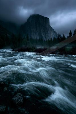 Turbulent river with white water rapids flows through a dark, misty mountain valley under heavy storm clouds, with a large rocky peak partially obscured by fog 