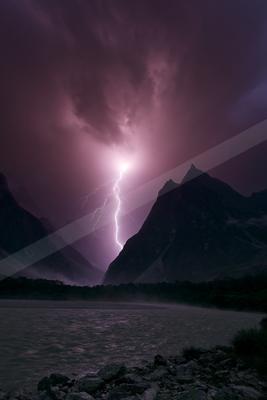 A powerful lightning bolt strikes from a dark purple stormy sky into a mountain valley above a rippling lake, with rocky shore in the foreground.