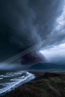 Moody dark storm clouds swirl above a rugged coastline where ocean waves crash onto a dark sand beach, with distant mountains under a brooding sky.
