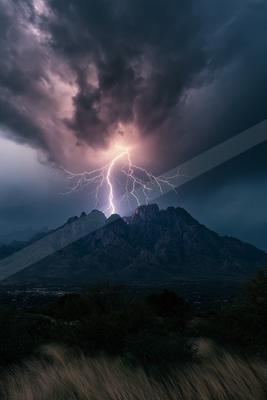A powerful lightning bolt strikes the peak of a rugged mountain range under a dramatic, dark stormy sky, with dry grass in the foreground.