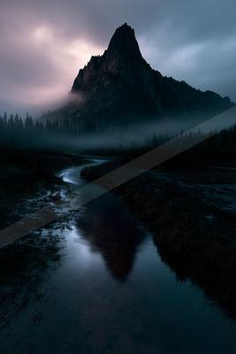 A sharp rocky mountain peak rises through thick fog above a dark coniferous forest, its silhouette perfectly reflected in a calm stream winding through a marshy