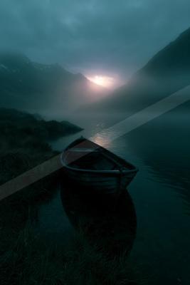 A wooden rowboat rests in shallow water on the edge of a misty mountain lake, with a glowing sun breaking through fog between dark peaks under a moody blue sky.