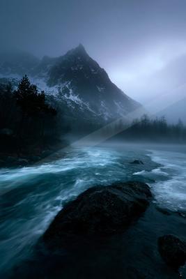 A turbulent river with white water rushes past dark rocks in the foreground, with a large snow-dusted mountain peak rising into thick mist and fog under a dark 