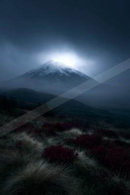 A snow-capped mountain peak emerges from thick fog with a bright light glowing from behind the summit, set against a dark moody sky, with purple and beige grass