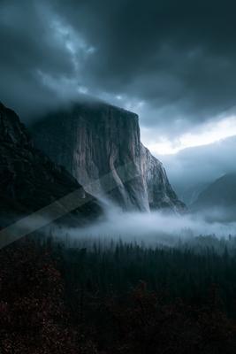 A massive rocky cliff face rises above a dense sea of fog in a forested mountain valley under dark, stormy clouds.