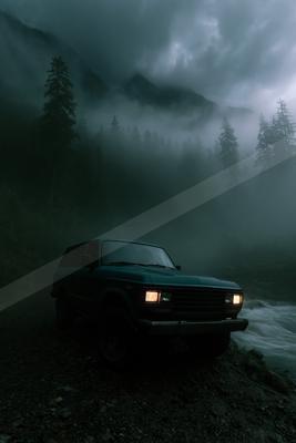 A dark green off-road vehicle with headlights on is parked on a rocky path beside a rushing river in a misty, forested mountain valley at dusk.