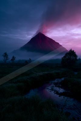A large volcanic mountain with its peak illuminated in red and purple hues rises above a layer of mist, under a dramatic twilight sky. In the foreground, a smal