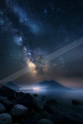 The Milky Way stretches across a starry night sky above a calm mountain lake and dark rocky foreground.