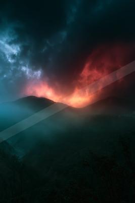 A dark mountain range is silhouetted against a dramatic sky filled with swirling clouds illuminated by intense red and orange hues.