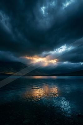 Moody landscape of a calm lake reflecting golden light from a break in dark storm clouds above distant mountains