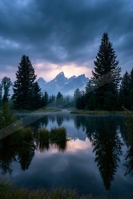 A serene lake reflects the silhouettes of tall evergreen trees and distant mountain peaks under a moody, cloudy sky during twilight.