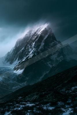 A towering, jagged mountain covered in snow and ice rises into dark, stormy clouds with mist swirling around its peak.