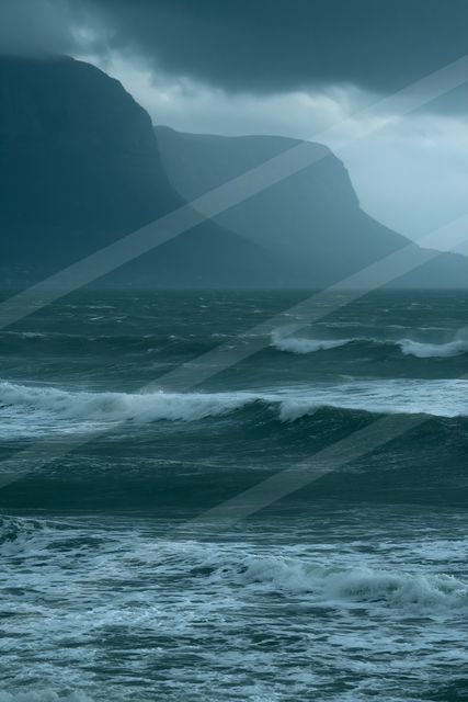 A moody seascape featuring turbulent ocean waves crashing against the shore, set against a backdrop of dark, imposing mountains under an overcast sky.