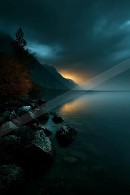 A rocky lakeshore with dark boulders and autumnal trees under a stormy sky, with a band of golden light breaking through the clouds and reflecting on the calm w