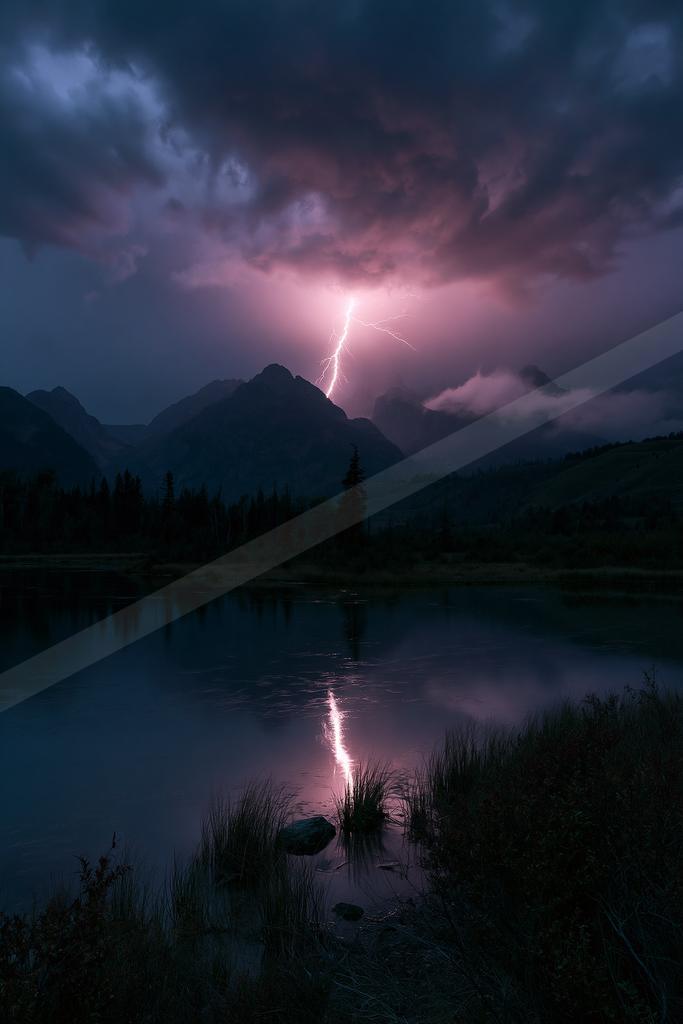 A dramatic lightning bolt strikes a mountain peak during a thunderstorm at night, its bright reflection visible on the calm surface of a foreground lake surroun