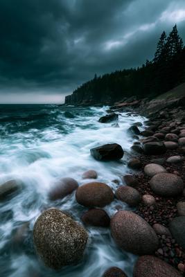 Powerful turquoise and white waves crash against a rocky shoreline under a dramatic, dark stormy sky, with a forested cliff in the background.