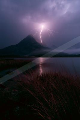 A bright lightning bolt strikes a mountain peak during a dramatic storm, its light reflecting on a calm lake in the foreground surrounded by dark reeds and rock