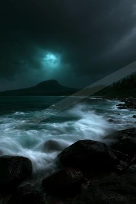 Powerful ocean waves crash against dark volcanic rocks in the foreground, with a turbulent sea and a distant mountain silhouetted under a dramatic stormy sky pi