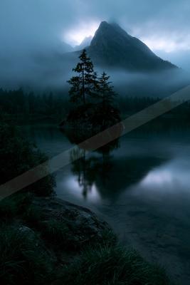 A serene mountain lake shrouded in thick fog, featuring a small rocky island with tall pine trees whose reflections are visible in the calm water, with a dramat
