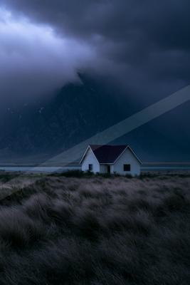 A solitary white house with a dark roof stands in a field of tall grass with a large mountain partially obscured by heavy storm clouds in the background.