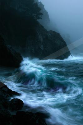 Powerful turquoise and white waves crash against dark rocky cliffs under thick fog, with silhouetted trees on the headland.