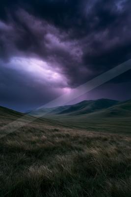 Moody landscape with dark purple storm clouds, visible lightning strike, and rolling green hills covered in dry grass under a dramatic sky.
