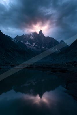 A jagged, snow-dusted mountain peak rises under brooding storm clouds with a bright light breaking through, perfectly mirrored in a still foreground lake.