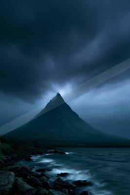 A sharp, dark mountain peak rises above a rocky lakeshore under heavy, moody storm clouds with a faint glow breaking through at the summit.