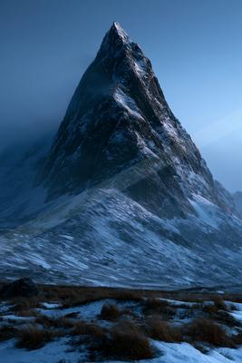 A sharp, snow-dusted mountain peak rises dramatically into a blue sky, with low clouds wrapping around its rocky ridges above a snowy foreground with dry grass.