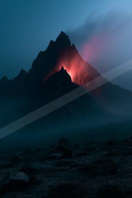 A jagged mountain peak glows with intense red light from volcanic activity against a dark blue night sky, with rocky foreground and atmospheric mist.
