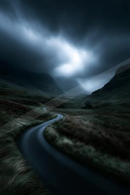 A dark winding road curves through a desolate grassy valley surrounded by large mountains under a dramatic, cloudy sky with a bright break in the clouds.