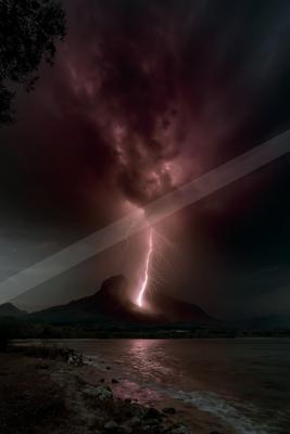 A powerful lightning bolt strikes a mountain peak during a thunderstorm, illuminating dark purple clouds and reflecting on the surface of a lake in the foregrou